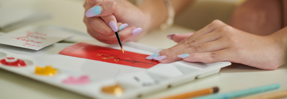 Person's hand holding a paint brush to hand-paint a card with paint supplies around.