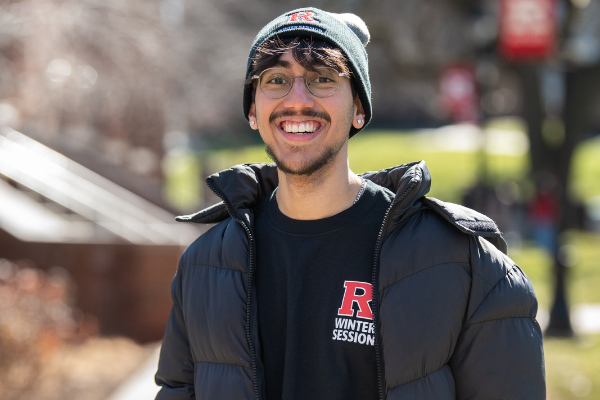 Current Rutgers student in a Winter Session hat and shirt with a jacket on, smiling on campus.