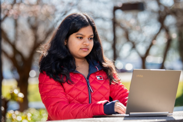 Student in a red jacket outside during winter typing on a laptop.