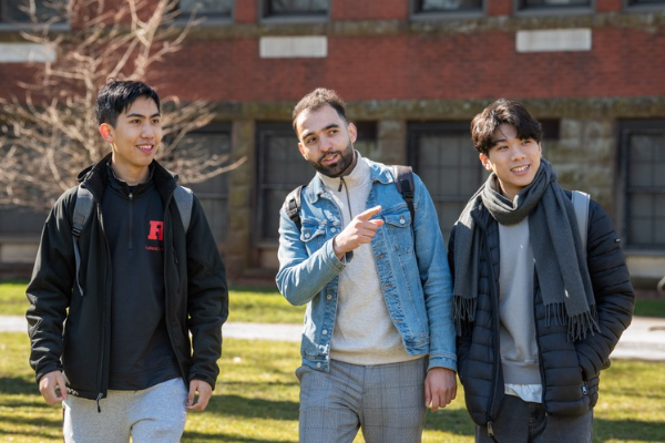 3 Rutgers students walking across campus in winter gear, one student in the middle is pointing toward the distance. These students are not International students, but Rutgers students.